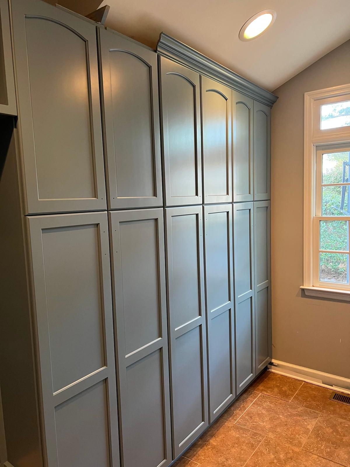 Tall pantry cabinets painted smooth gray-blue with crown molding beside a white-trimmed window