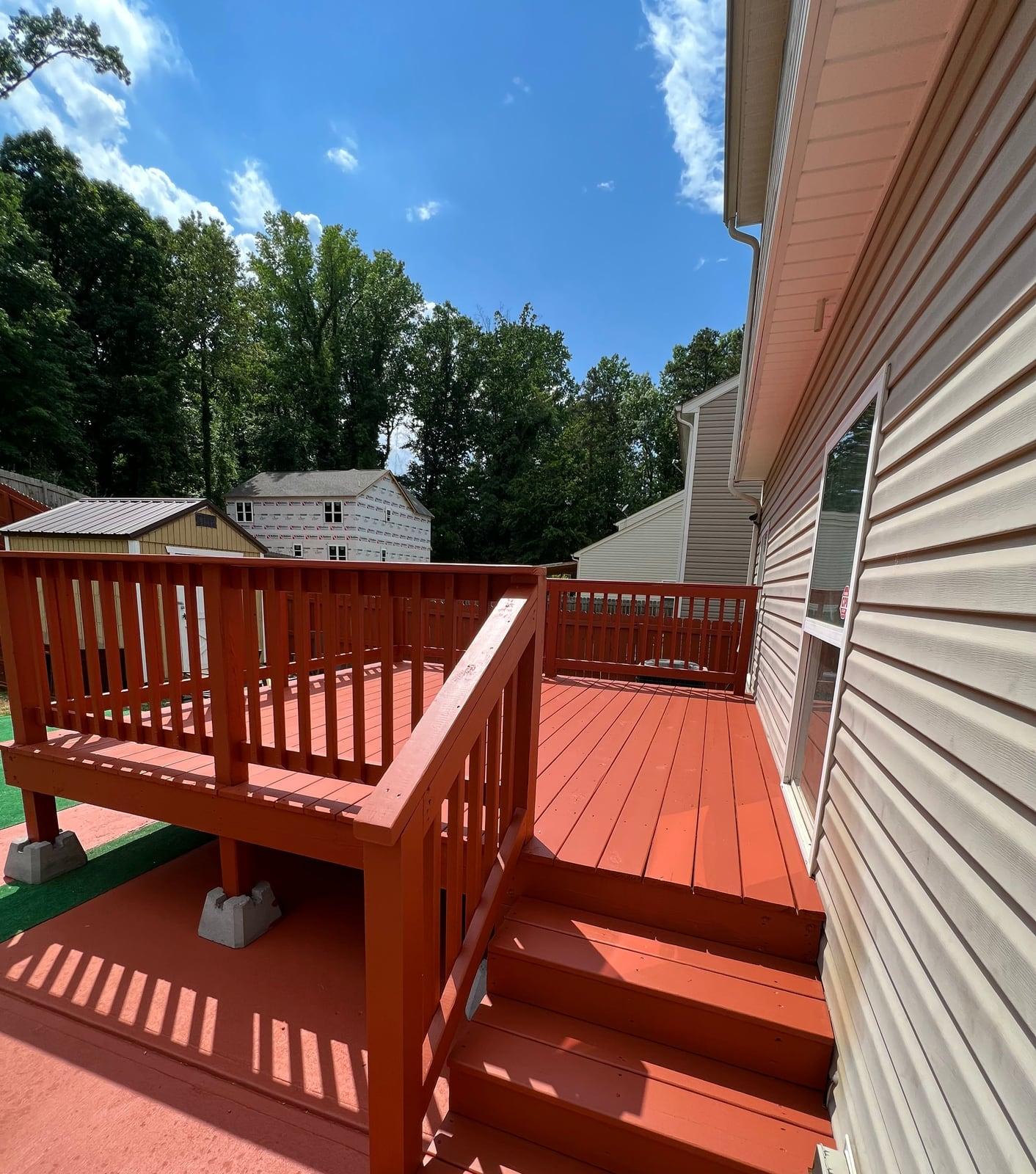 Freshly finished red-brown deck with railing and steps beside a house under bright blue sky